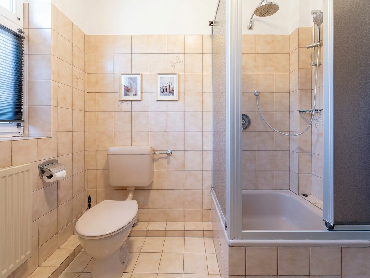 Kleines Badezimmer mit Toilette, Duschkabine, zwei gerahmten Bildern an der gefliesten Wand und einem Fenster mit Jalousie