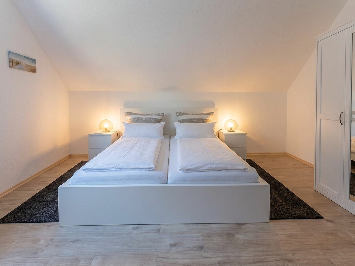 Schlafzimmer mit Doppelbett auf dunklem Teppich, zwei Nachttischen mit Lampen und weißem Kleiderschrank unter schräger Decke