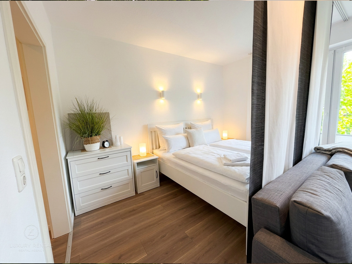 Schlafzimmer der Ferienwohnung Etta in Dangast mit Doppelbett, Nachttisch, Kommode mit Pflanze und Wecker sowie grauem Sofa am Fenster