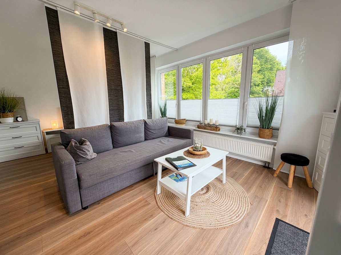 Helles Wohnzimmer der Ferienwohnung Etta mit grauem Sofa, weißem Couchtisch auf rundem Teppich vor großen Fenstern mit Blick ins Grüne